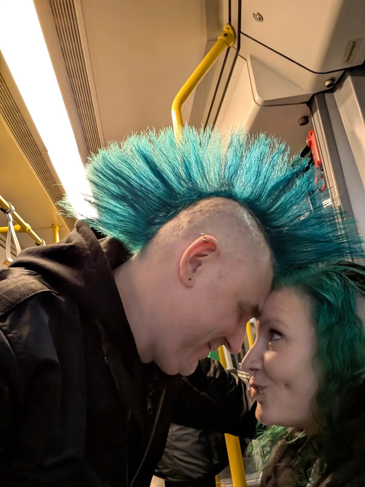 Mann mit hohem türkisem Iro und Frau mit grünen Haaren, Stirn an Stirn in der Berliner Tram