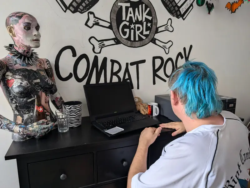 Person mit blauen Haaren am Laptop vor einem Combat Rock Wandbild
